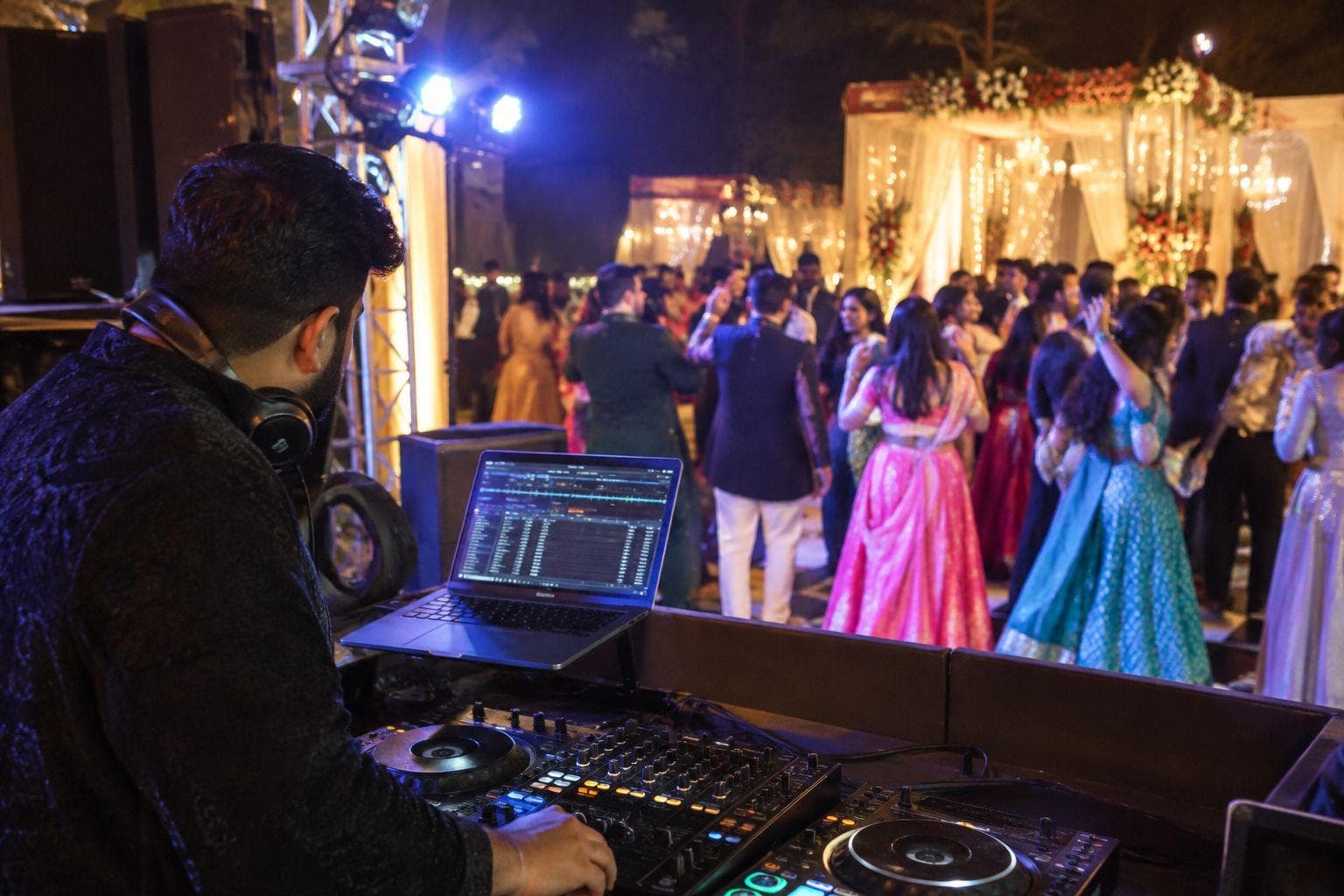 A DJ with headphones at the decks watching guests in colourful Indian wedding outfits dance beneath a lit floral mandap.