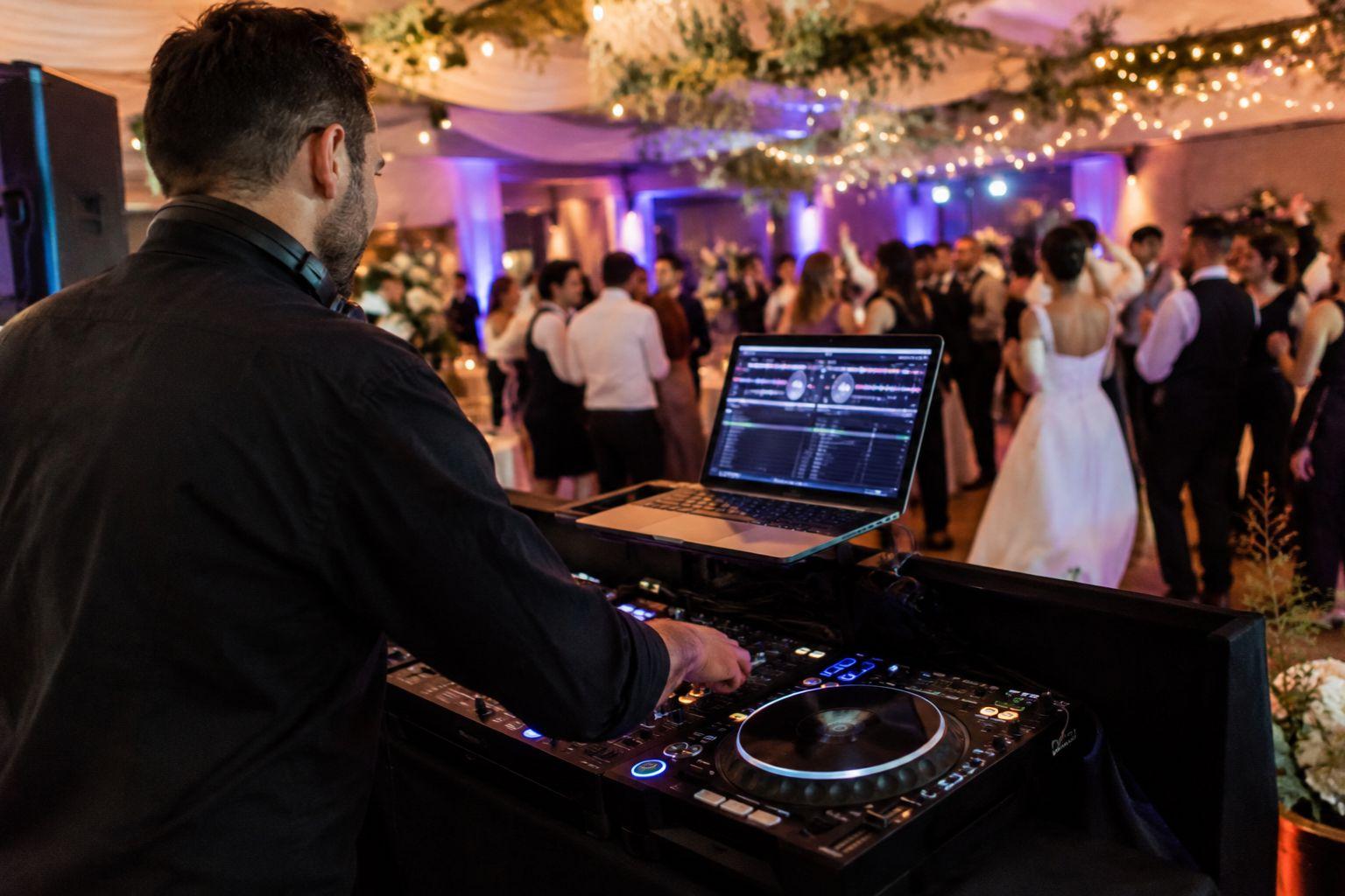 A wedding DJ at the decks watching a full dancefloor, with the couple dancing under festoon lights at a reception.