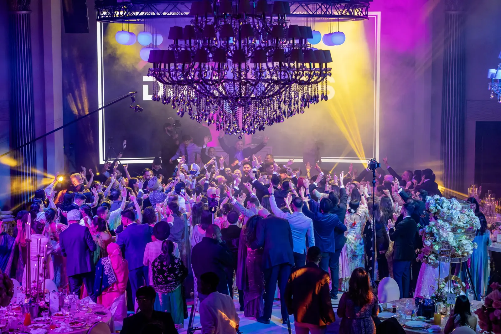 A packed wedding dancefloor beneath blue and purple stage beams at Dallas Burston Polo Club