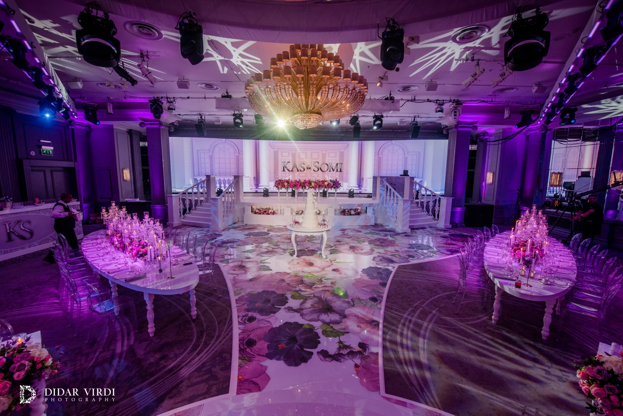 The room in violet light Purple lighting above the floral walkway, candlelit tables and white wedding stage at Grosvenor House Ballroom.