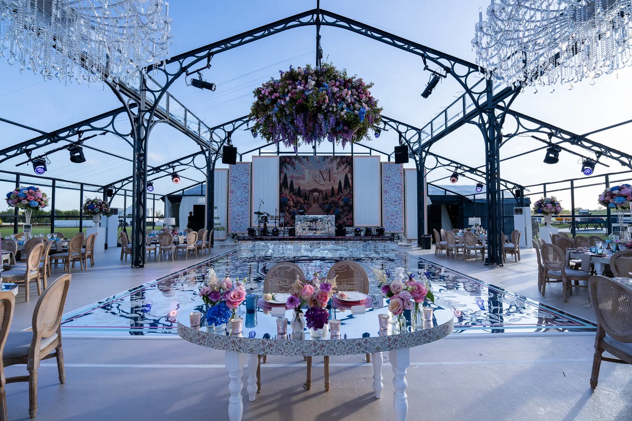 The sweetheart table at the edge of the mirror dancefloor, the wisteria floral chandelier hanging over the floor and the stage beyond, Jeevan and Megha's wedding in the glass marquee at Dallas Burston Polo Club, Kudos Music