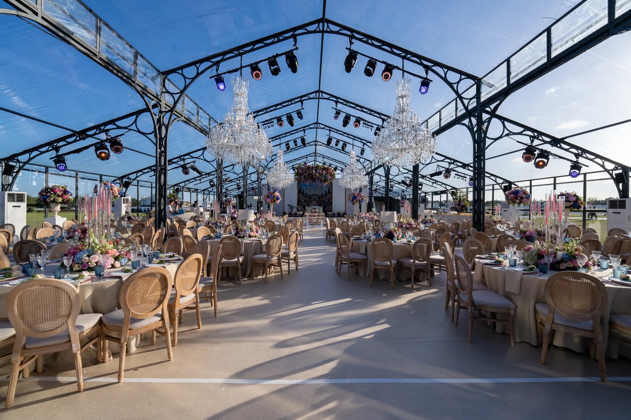 Round tables with wildflower centrepieces and pink taper candles down the length of the glass marquee, two crystal chandeliers and the rig on the frame above, Jeevan and Megha's reception at Dallas Burston, Kudos Music