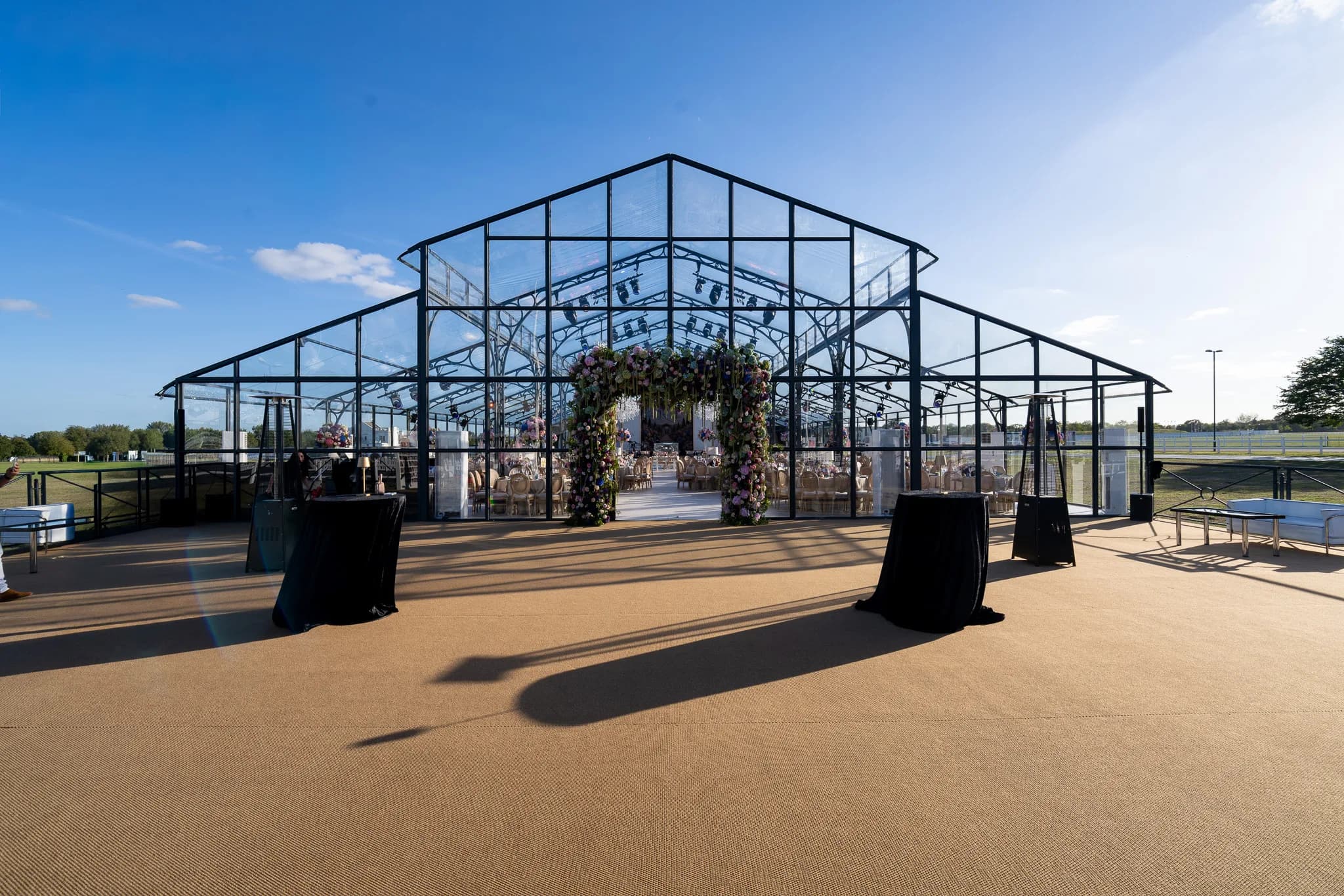 The front of the glass marquee with a floral arch at its door and poseur tables on the coir forecourt, Jeevan and Megha's wedding at Dallas Burston Polo Club, Kudos Music