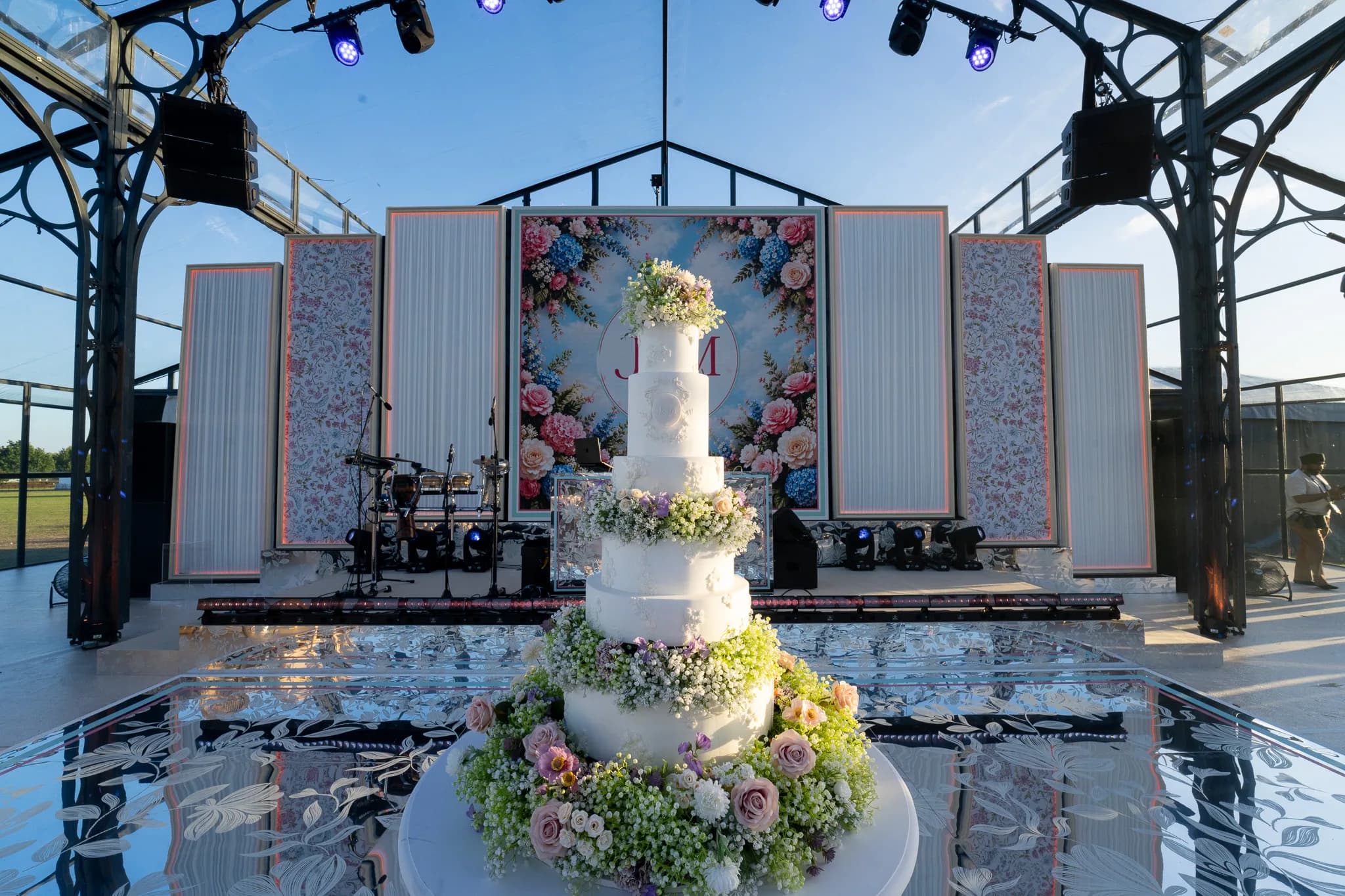 The five tier wedding cake by Maya Dahlia Cakes dressed in fresh roses and hydrangea on the mirror floor, the floral monogram on the LED screen behind it, Jeevan and Megha's reception at Dallas Burston, Kudos Music