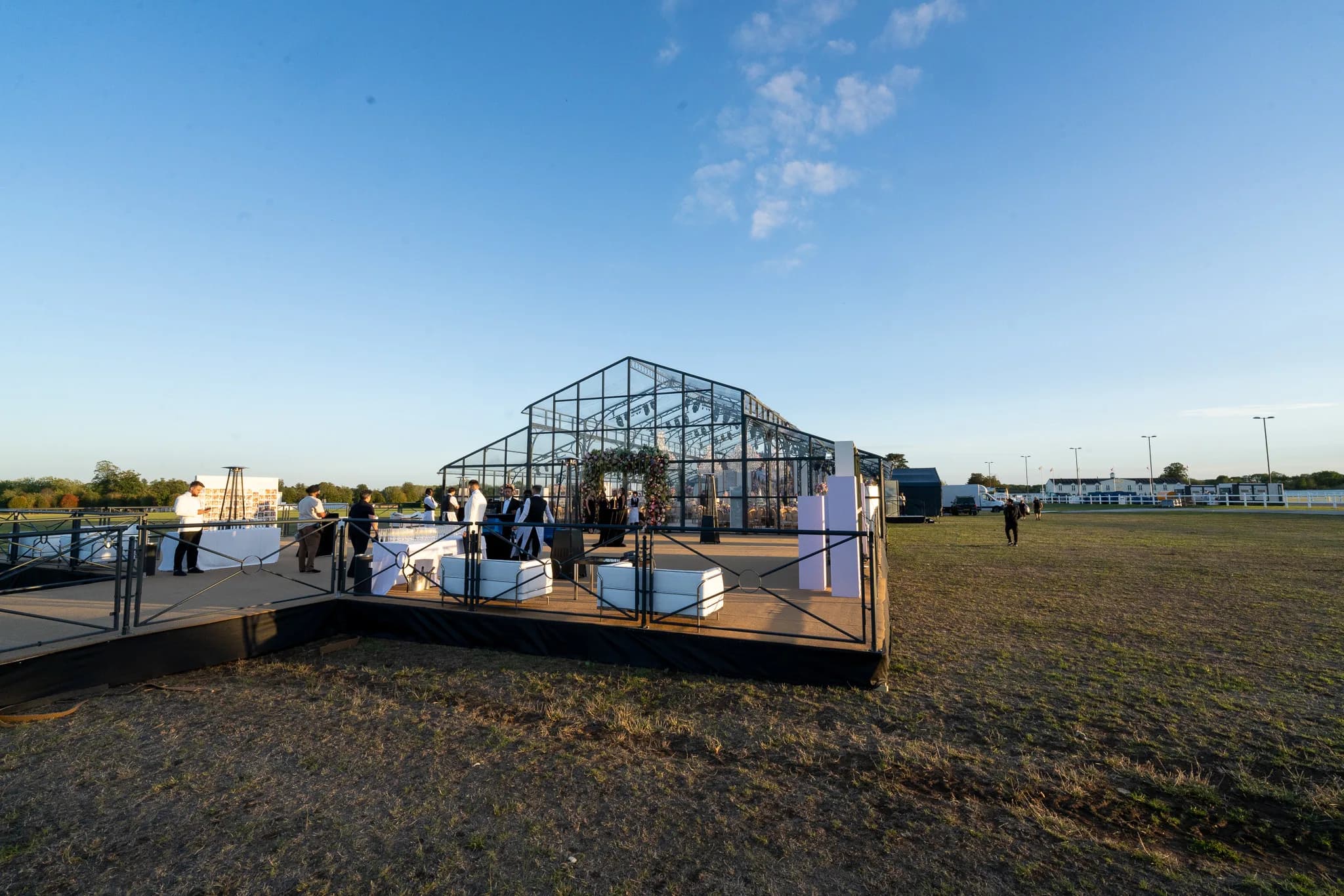 The deck outside the glass marquee at golden hour, white lounge seating and staff ready for the drinks reception on the polo grounds at Dallas Burston, Jeevan and Megha's wedding, Kudos Music