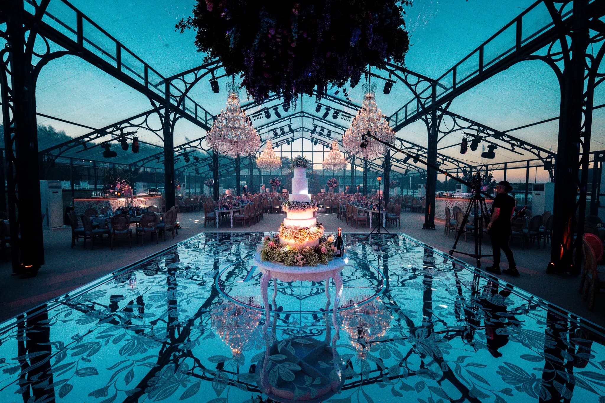 Blue hour inside the glass marquee: the cake lit on the mirror floor, the chandeliers warm above, the floral cloud over the floor and the film crew at work, Jeevan and Megha's reception at Dallas Burston, Kudos Music