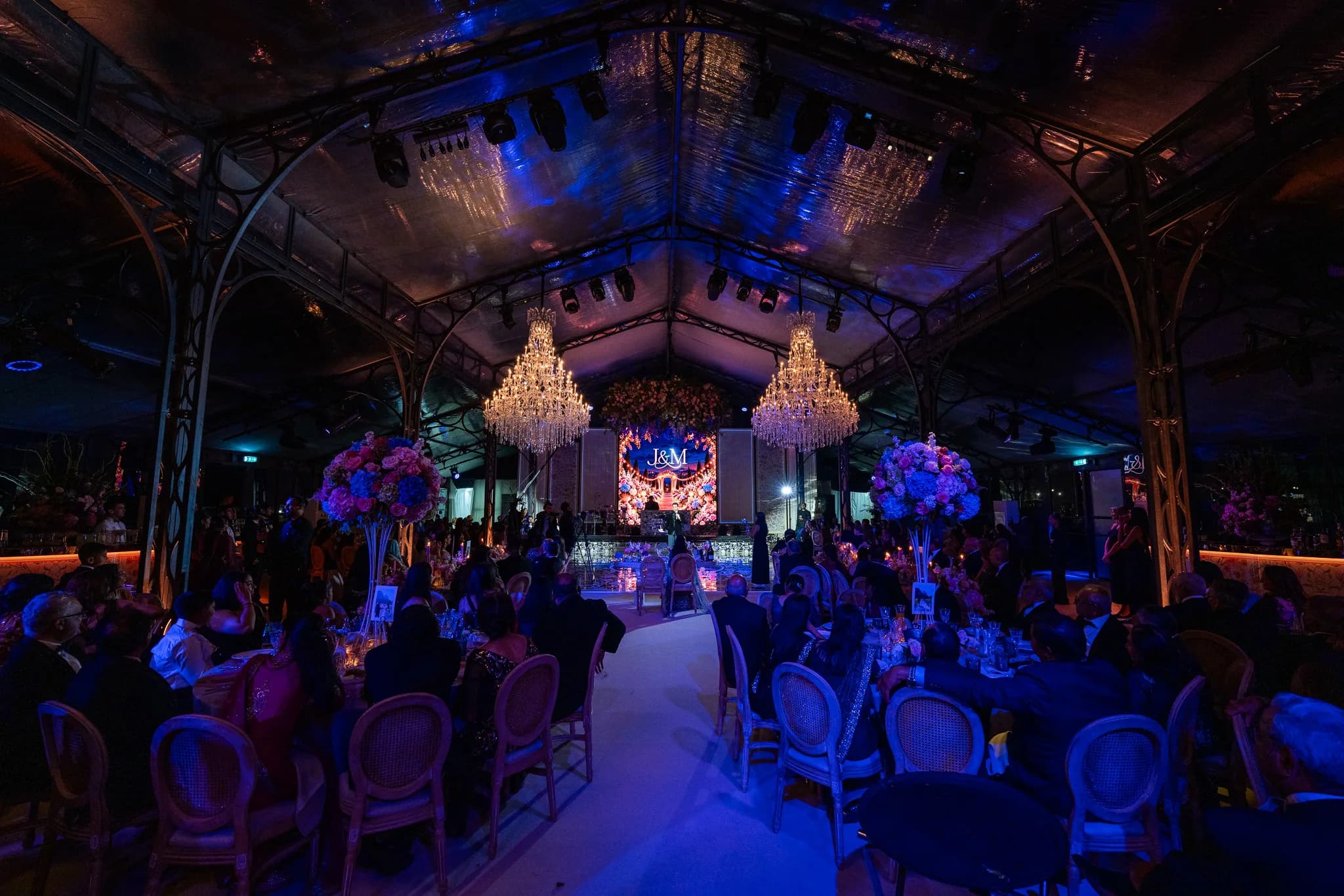 Dinner under blue light, tall pink and blue floral centrepieces on the tables and Jeevan and Megha's monogram on the screen between the chandeliers, Dallas Burston Polo Club, Kudos Music