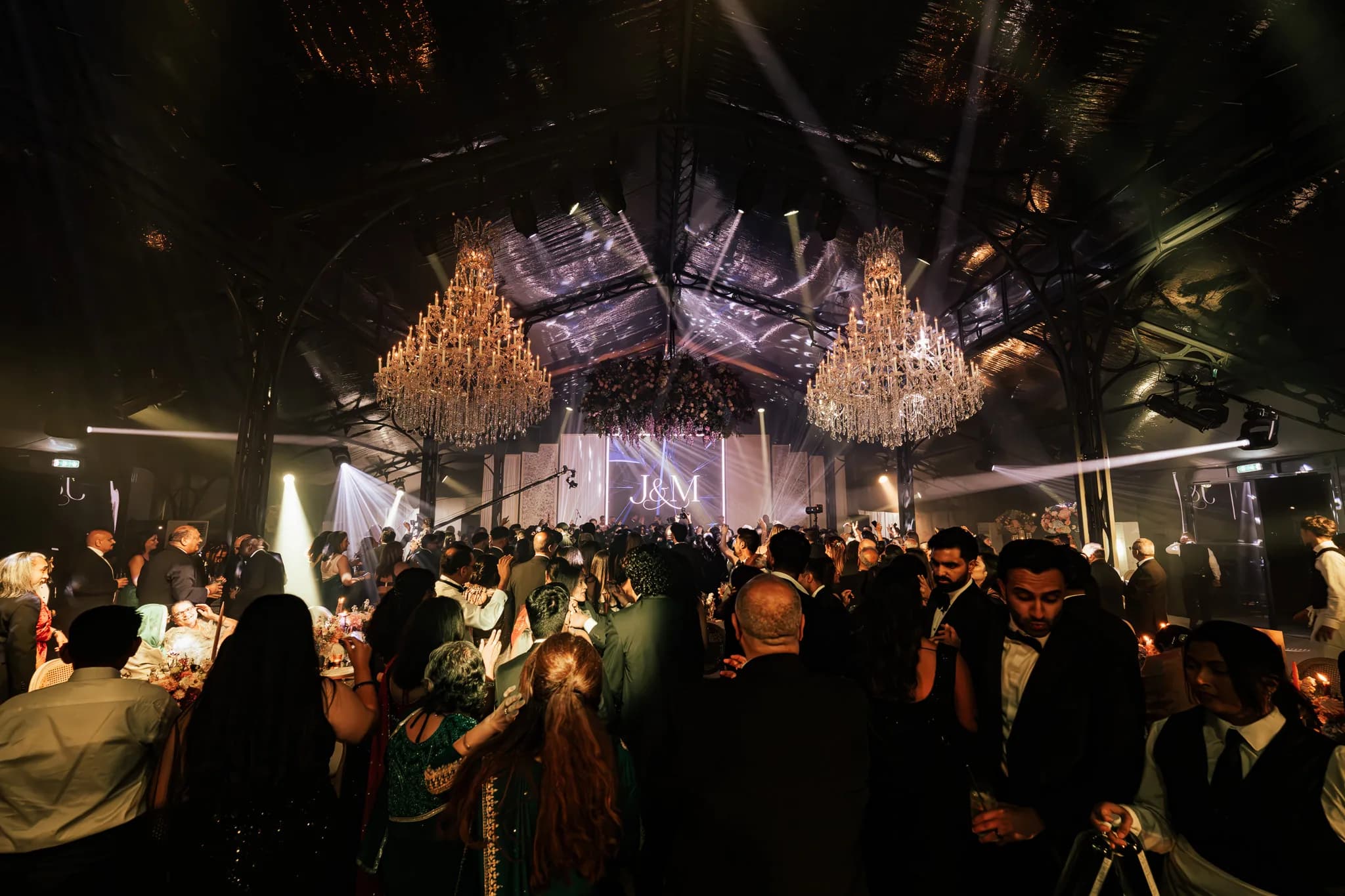 The party from the back of the room, beams through haze, the chandeliers lit warm and the monogram on the screen, Jeevan and Megha's reception in the glass marquee at Dallas Burston, Kudos Music