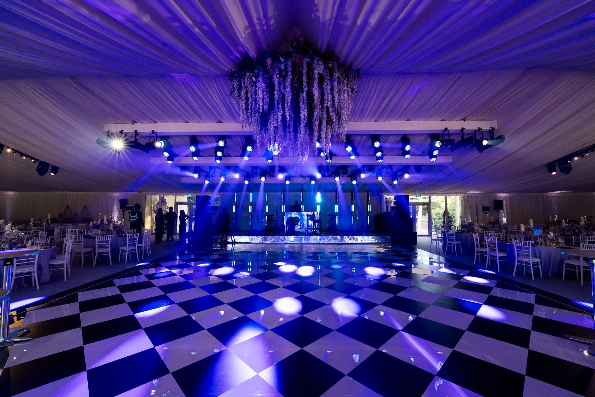 The mirror dancefloor lit blue with the dinner tables set around it and florals overhead, Pennyhill Park marquee wedding, Kudos Music