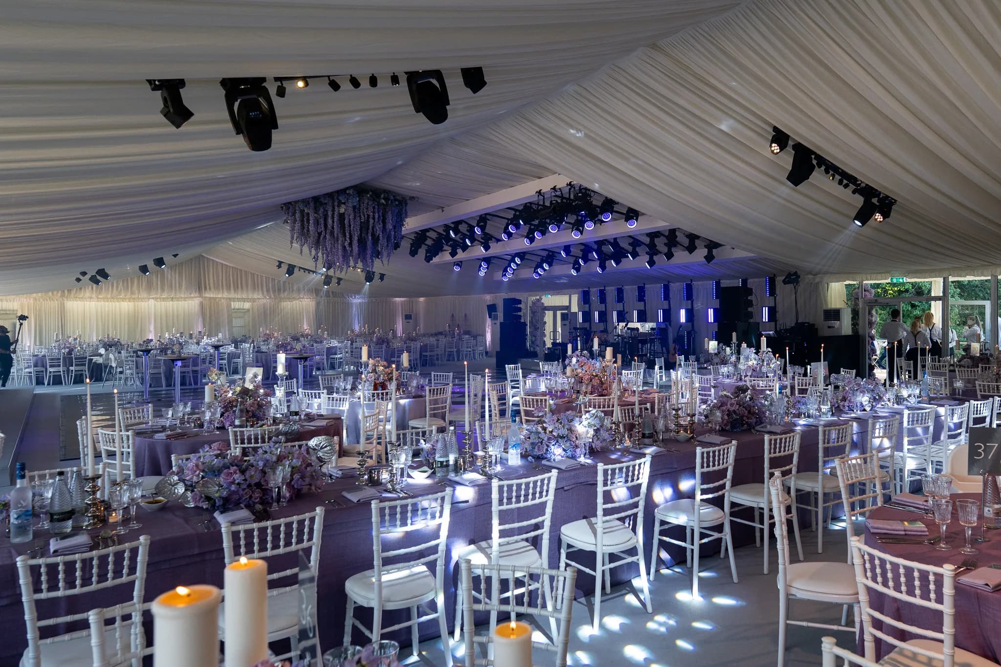 White chiavari chairs and purple floral centrepieces on the round tables, the marquee uplit blue, Lakhita and Satnaam's wedding at Pennyhill Park, Kudos Music