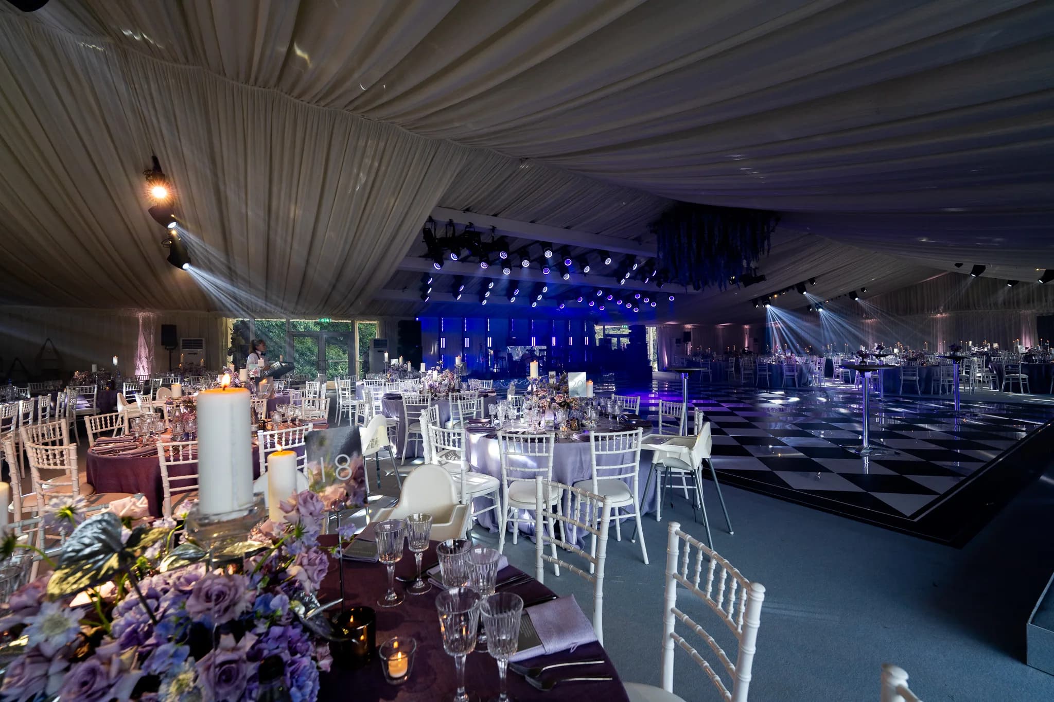 Purple florals on the tables, blue light on the stage and the draped ceiling of the marquee by Mahood Marquees at Pennyhill Park, Kudos Music