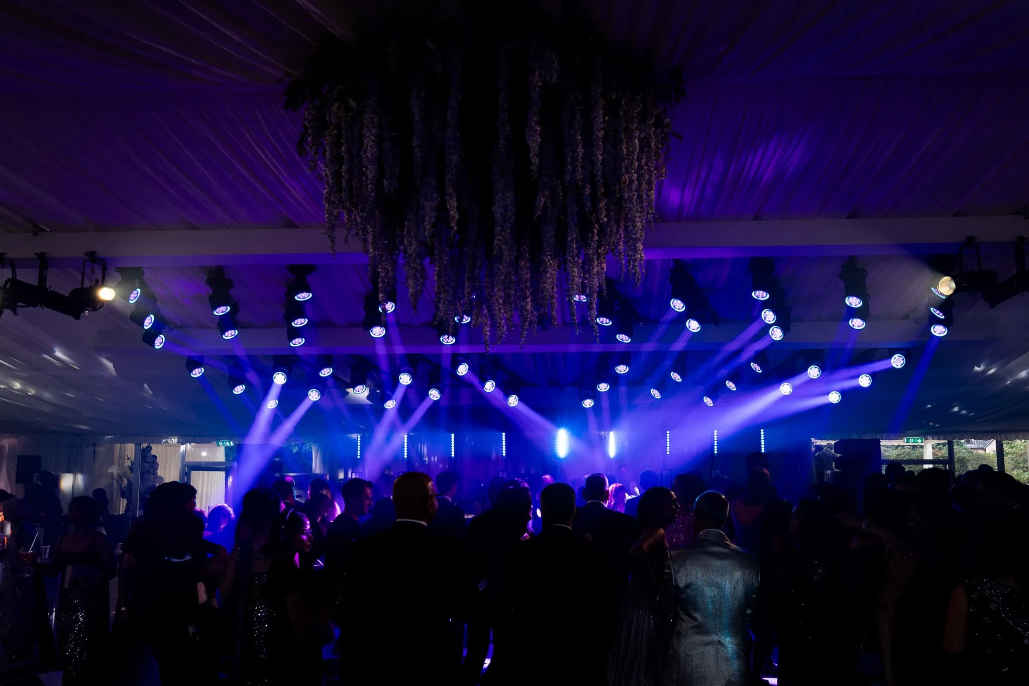 The dancefloor full under purple and blue light with florals overhead at Lakhita and Satnaam's reception, Pennyhill Park, Kudos Music