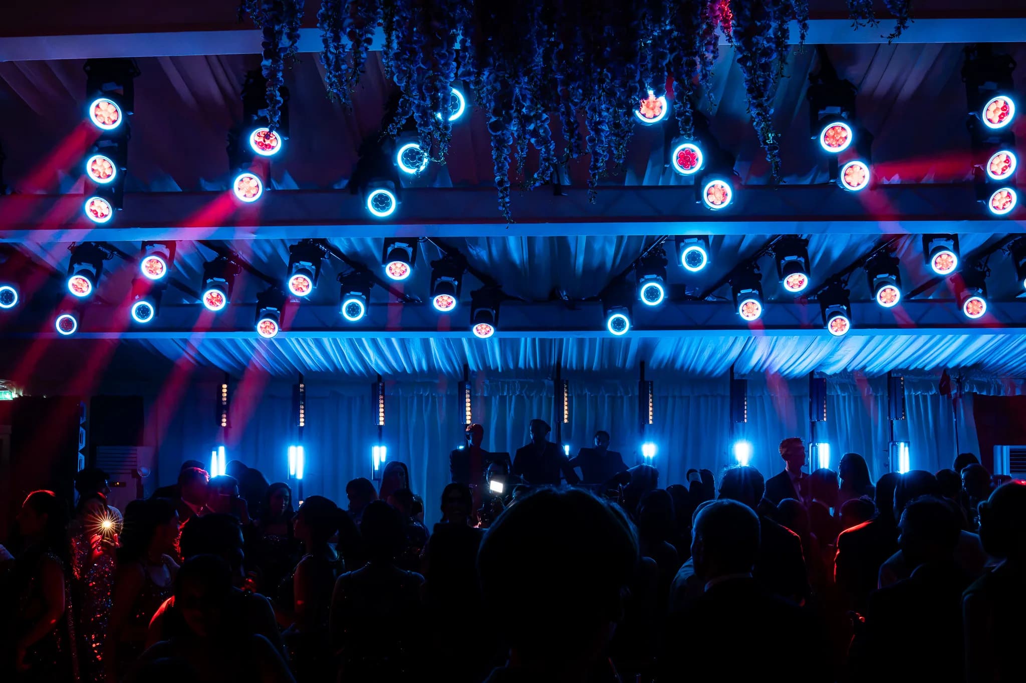 Circular light fixtures and hanging vines above the crowd, lit blue and red, at Lakhita and Satnaam's Pennyhill Park reception, Kudos Music