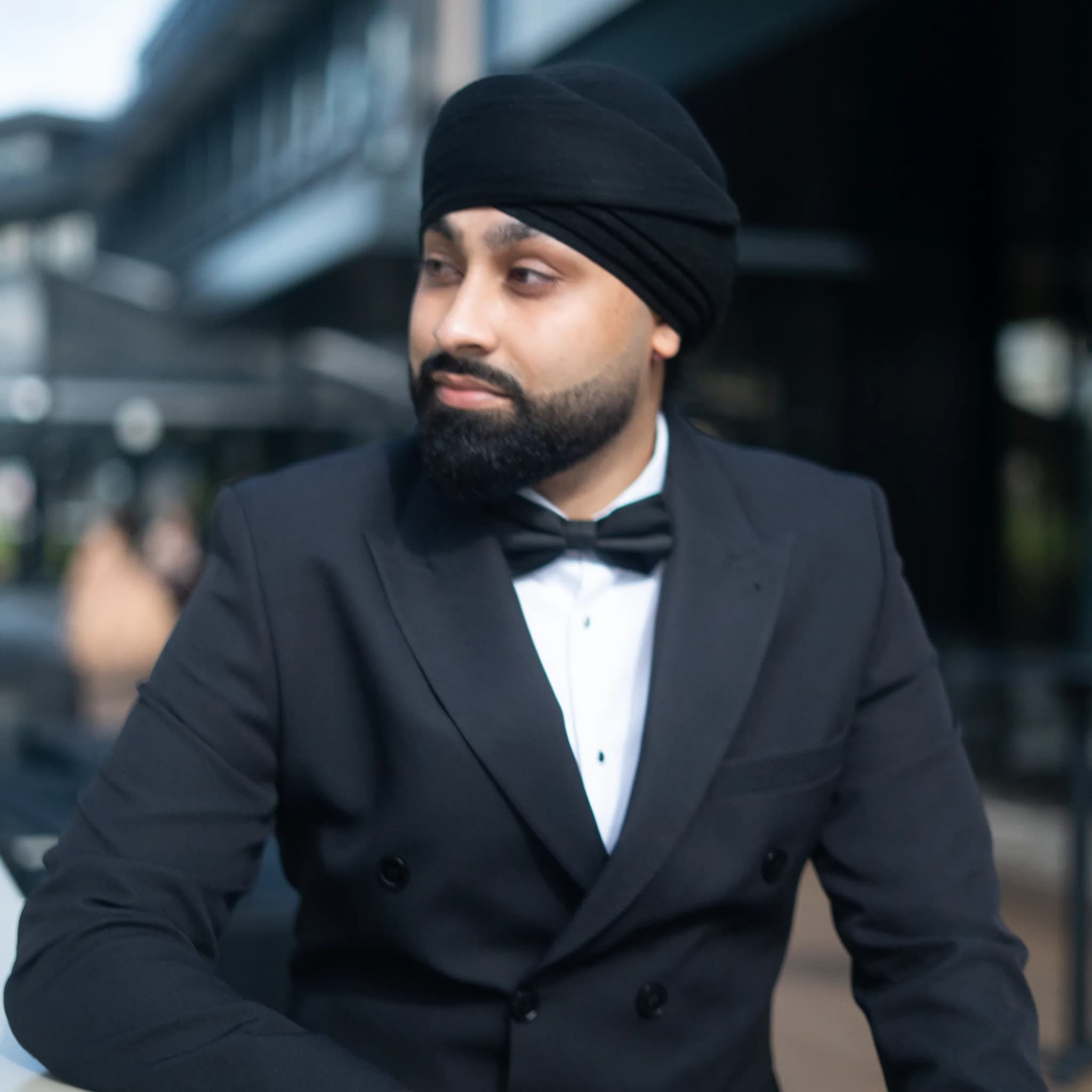DJ Harj Matharu of Kudos Music in a black tuxedo and turban, looking over his shoulder on a city street