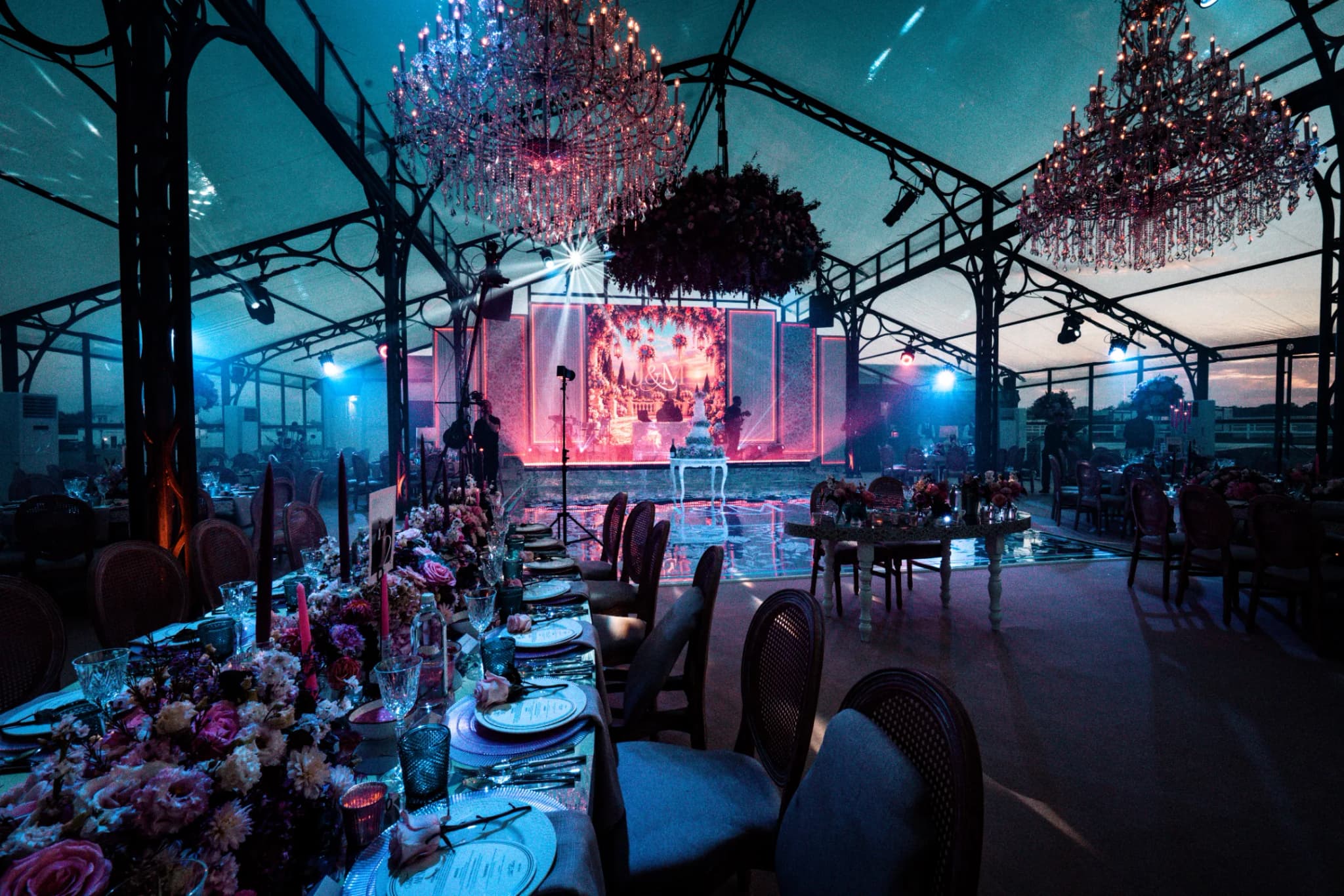 A long dining table dressed in pink and coral florals under two chandeliers lit red, looking across to the cake, the mirror floor and the screen, Jeevan and Megha's reception in the glass marquee at Dallas Burston Polo Club, Kudos Music