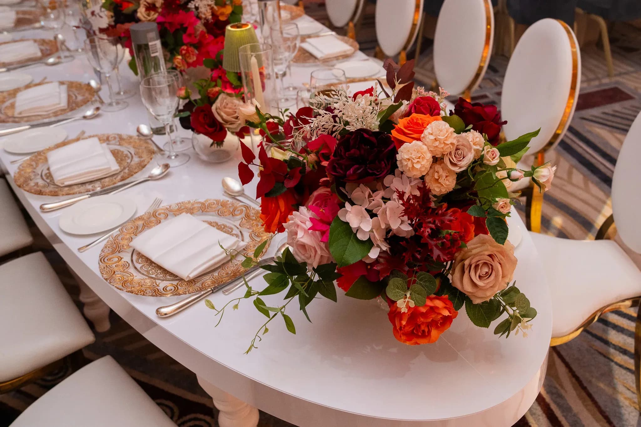 A place at the celebration | Royal Lancaster London | Kudos Music Gold-rimmed place settings and red flowers follow a curved white table at Pooja and Gary’s wedding.