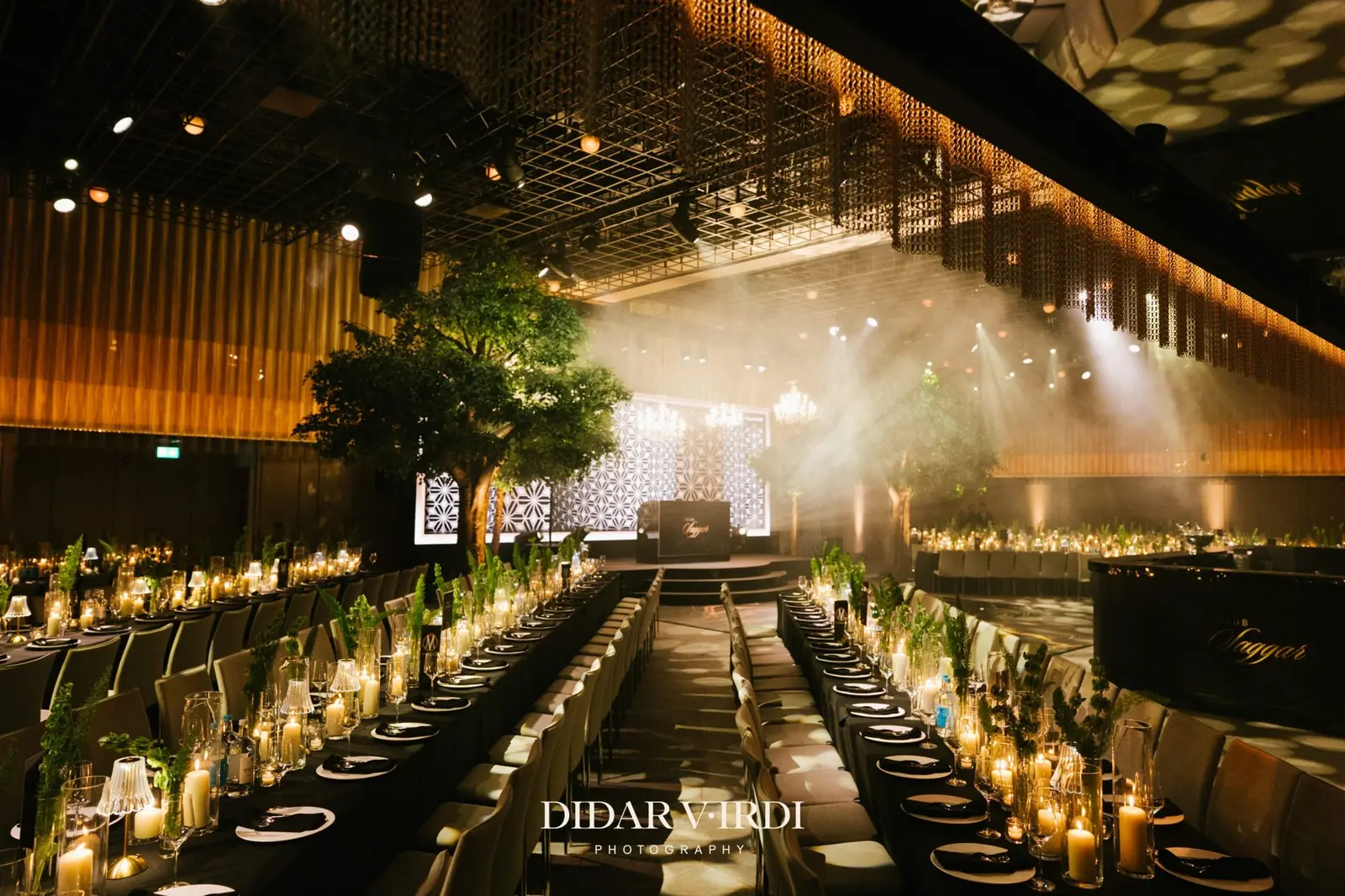 Central aisle between banquet tables facing an LED stage and chandeliers at The Londoner